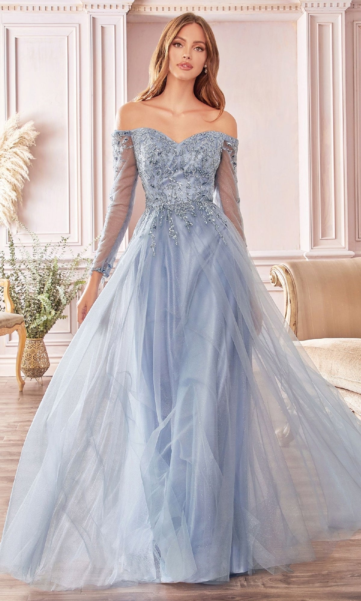 A woman wears the Ladivine CD0172, a light blue off-the-shoulder long prom dress with long sleeves and floral embellishments, while standing in an elegant, softly lit room with classic decor.