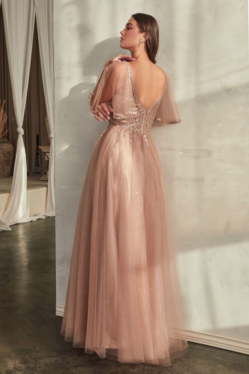 A woman in the Ladivine CD0175 Bell-Sleeve Glitter Formal Dress, a floor-length blush gown with sheer bell sleeves and sequin embellishments, stands facing a wall in a softly lit room.