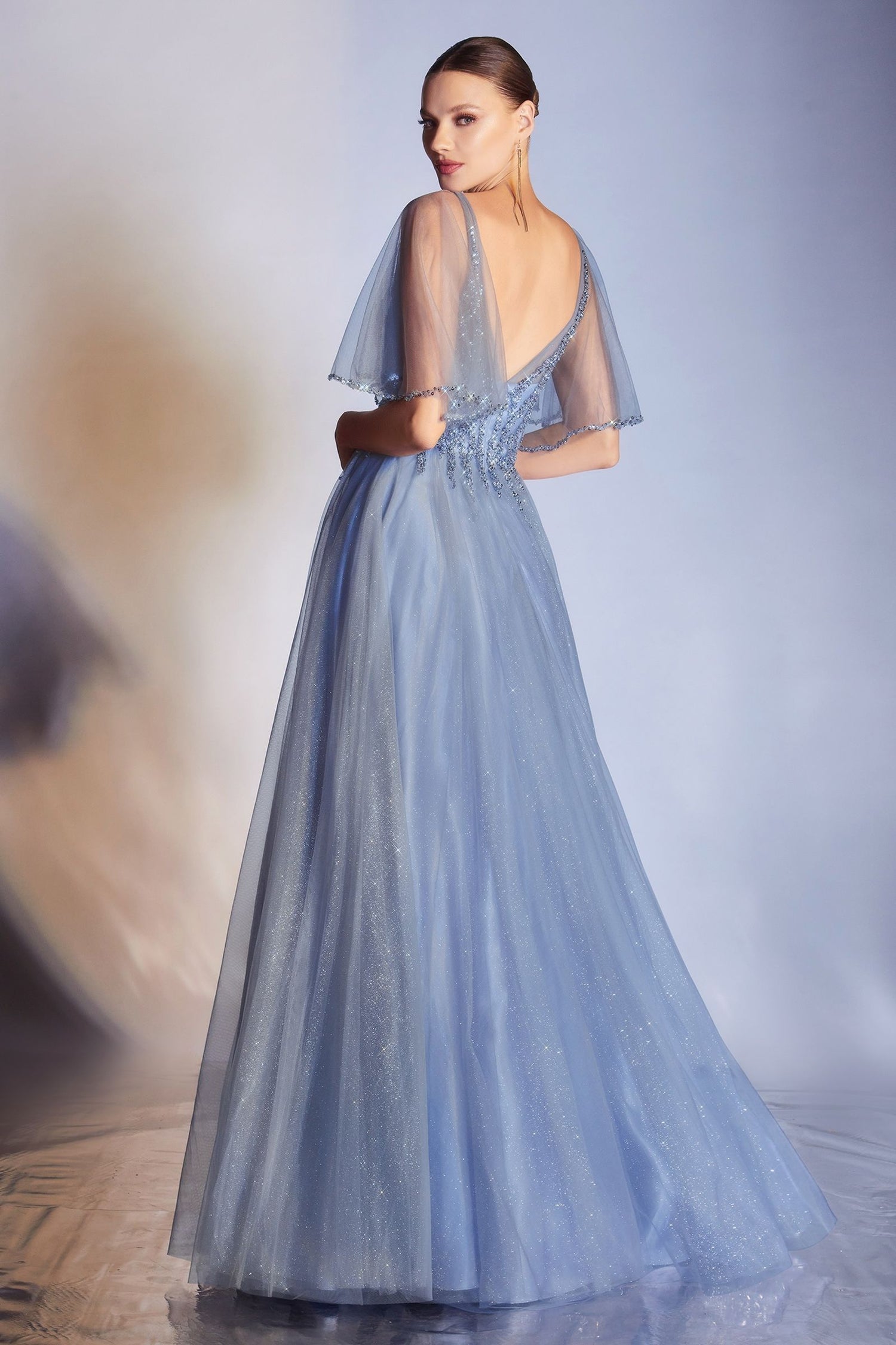 A woman faces away, wearing the Ladivine CD0175 Bell-Sleeve Glitter Formal Dress—a floor-length light blue gown with sheer short bell sleeves, beaded details, v-neckline, and a deep V-back—set against a softly lit background.