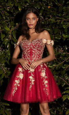 A woman wearing the Off-Shoulder Short Floral Homecoming Dress CD0194, featuring gold 3-D floral embellishments, stands before lush green leafy foliage.