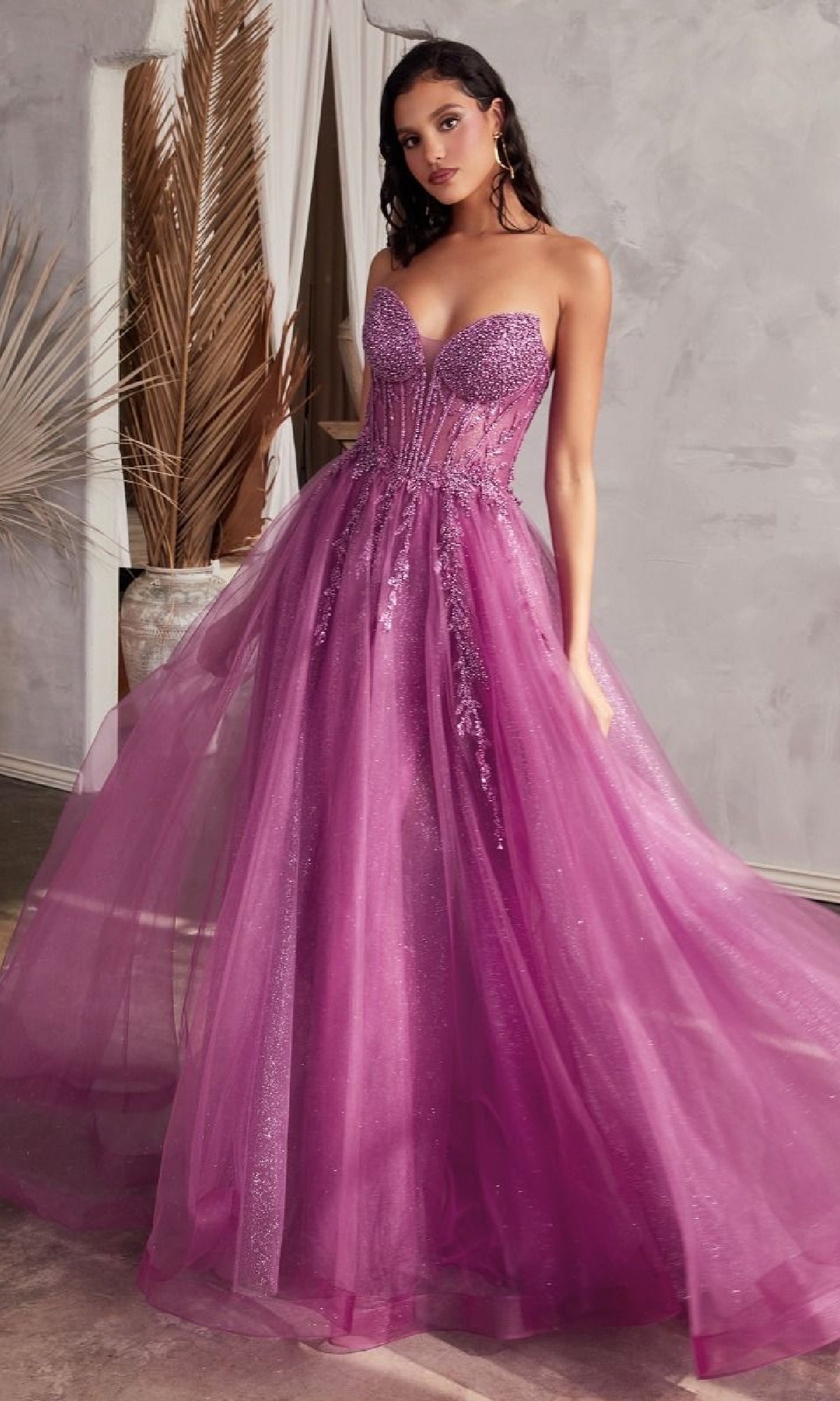 A woman stands indoors wearing the Strapless Sheer-Corset Long Prom Ball Gown CD0230—an elegant, floor-length purple tulle dress with a fitted, embellished bodice and flowing skirt.