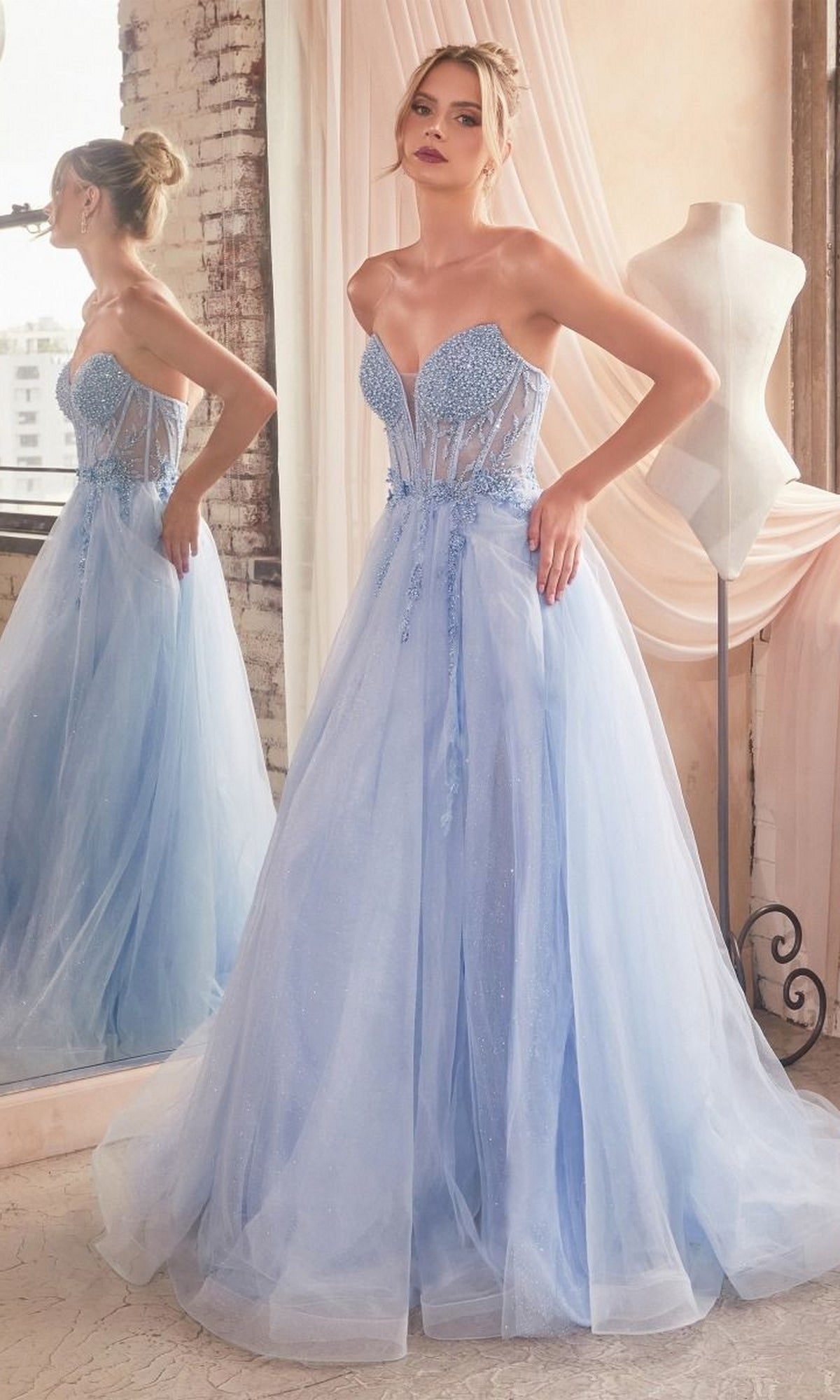 A woman stands before a mirror in the Strapless Sheer-Corset Long Prom Ball Gown CD0230, a light blue dress with a beaded sheer bodice, beside a dress form in a gently lit room.