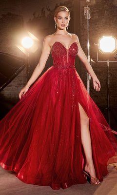A woman wearing the Strapless Sheer-Corset Long Prom Ball Gown CD0230, a strapless, floor-length red gown with a high slit and beaded bodice, stands before studio lights against a dark background.