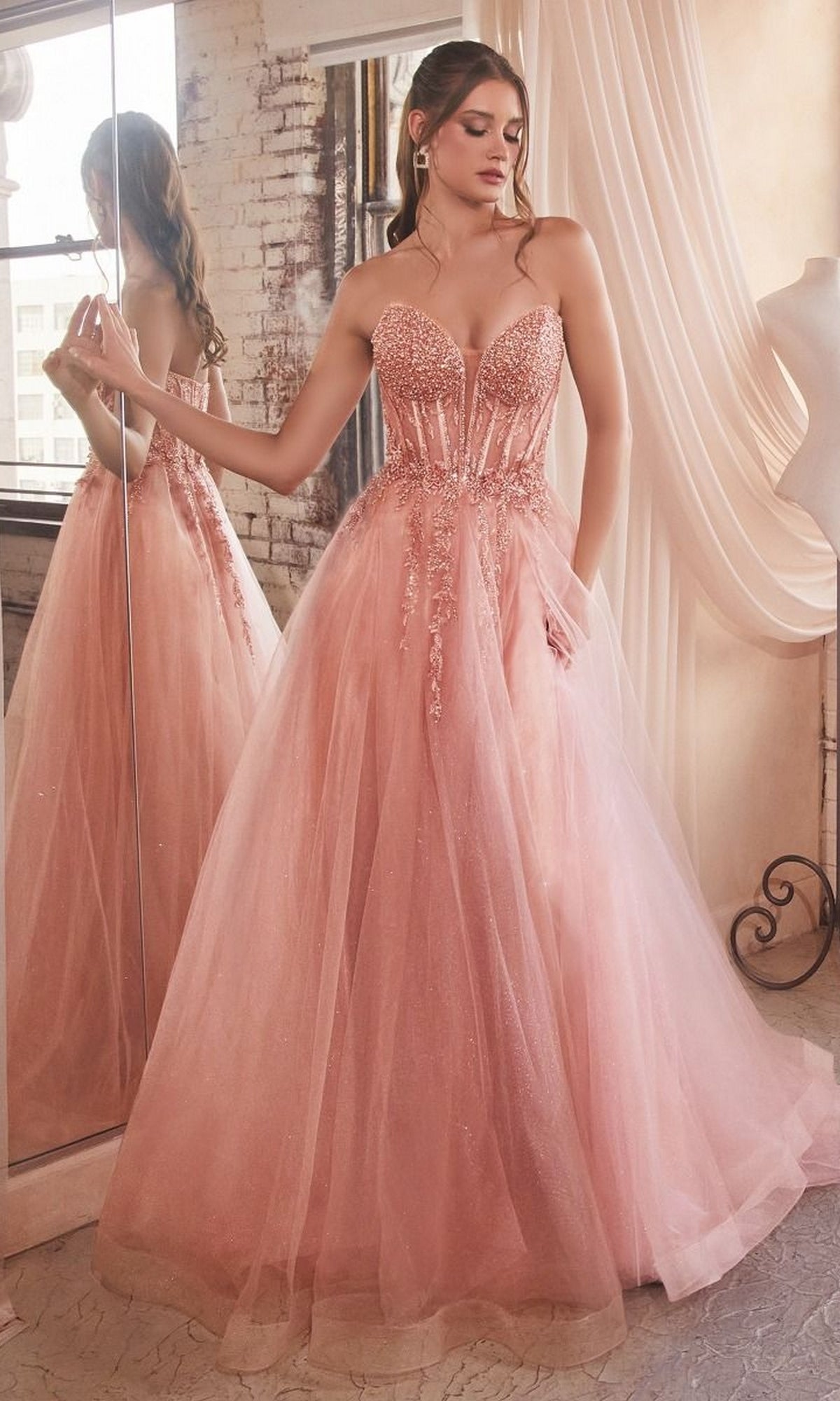 A woman in the Strapless Sheer-Corset Long Prom Ball Gown CD0230, featuring a fitted embellished bodice and full glitter tulle skirt, stands in a softly lit room with draped curtains and a large mirror.