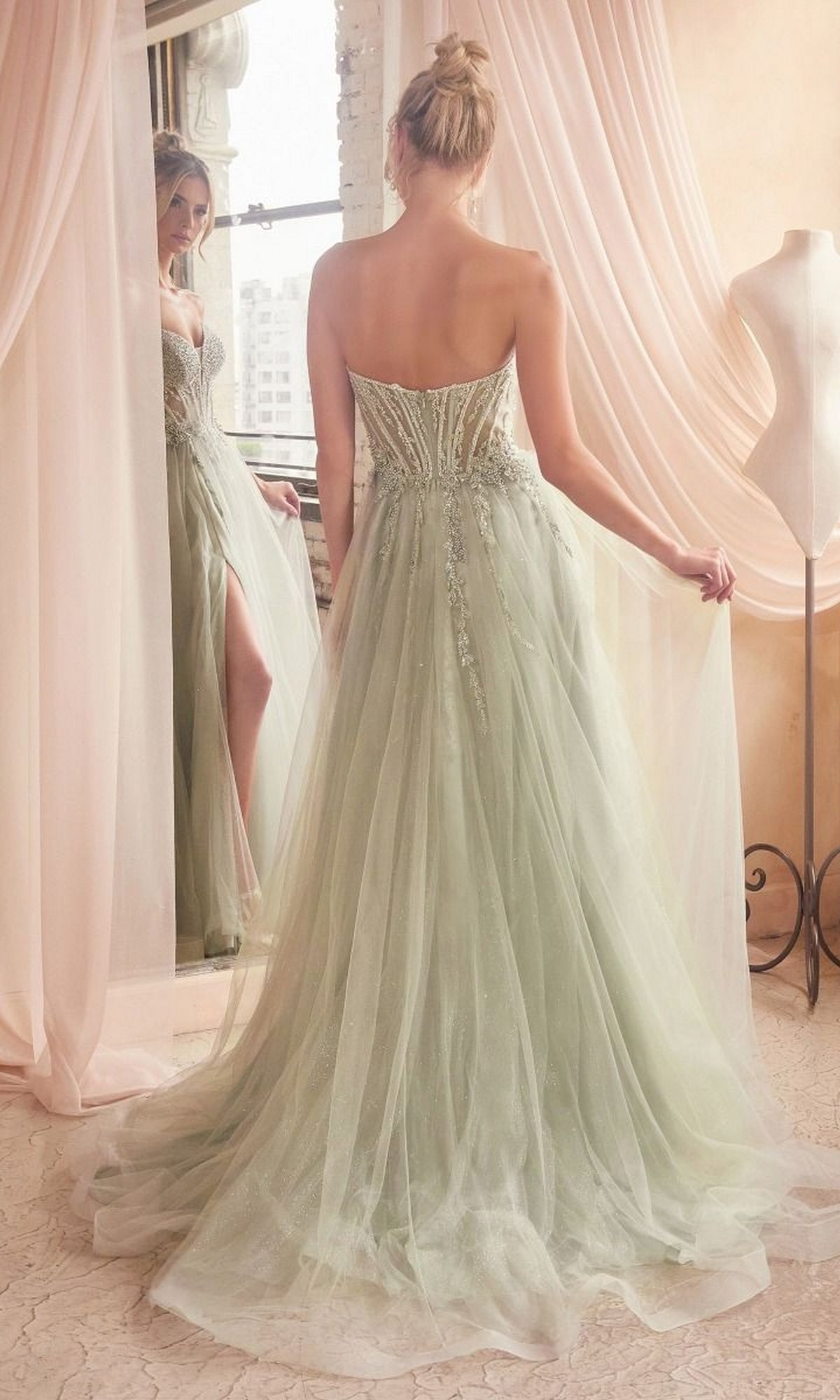 A woman in the Strapless Sheer-Corset Long Prom Ball Gown CD0230, made of light green tulle with beading, stands before pink curtains, gazing at her reflection. A dress form nearby displays a glitter tulle evening dress.