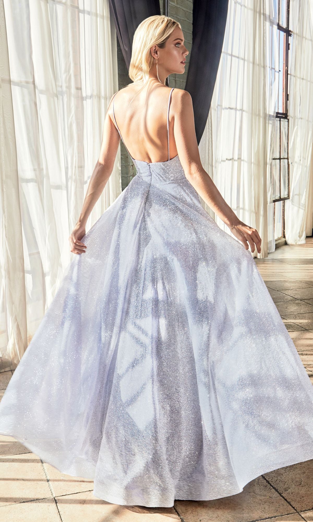 A woman stands indoors by large windows, wearing the Light Blue Glitter Long A-Line Prom Dress CD205 featuring a sleeveless design, shimmering fabric, low back, and full formal skirt.