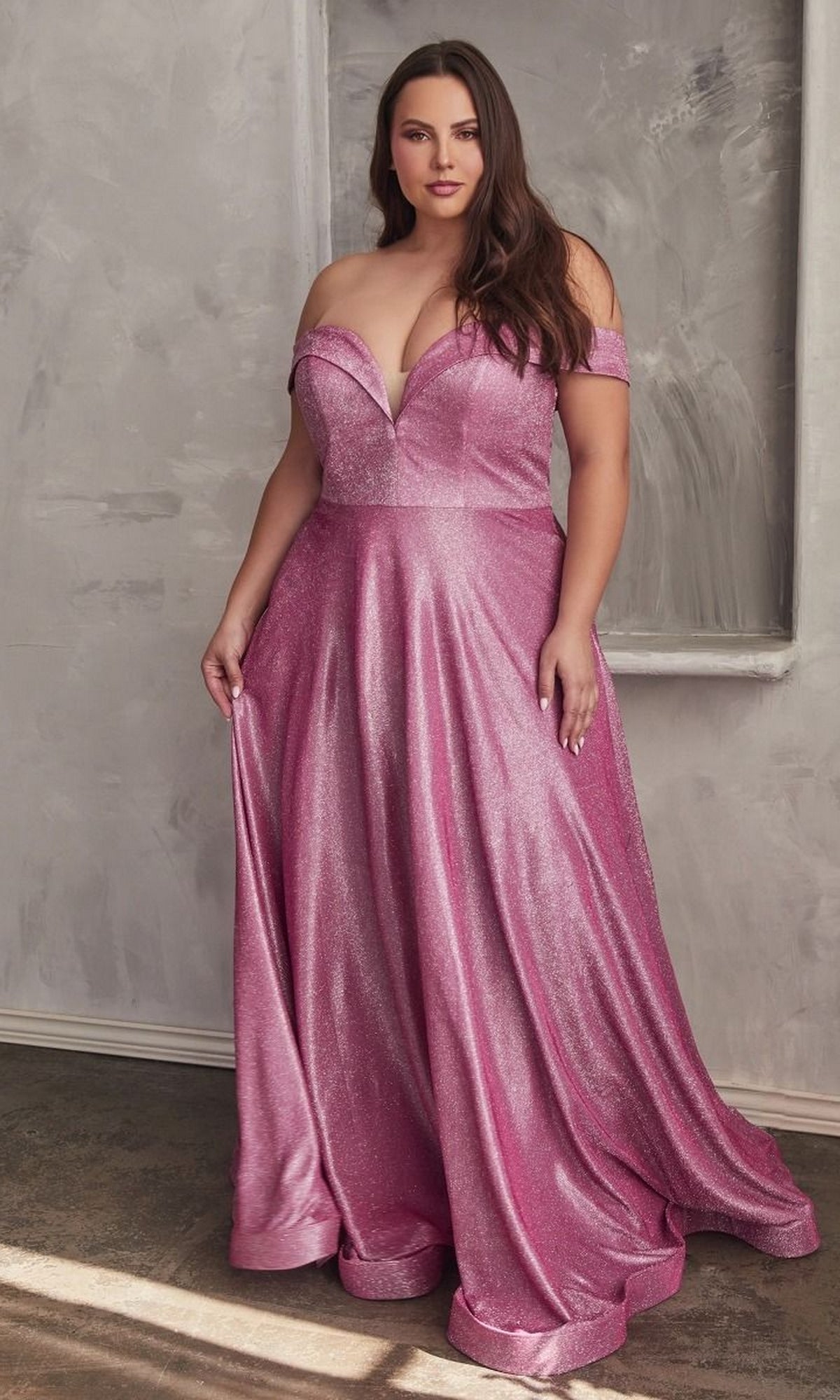 A woman stands indoors wearing the Plus-Size Long Metallic Lace-Up Prom Dress CD210C, featuring a shiny off-the-shoulder design with a fitted bodice and flowing skirt, ideal for formal occasions.
