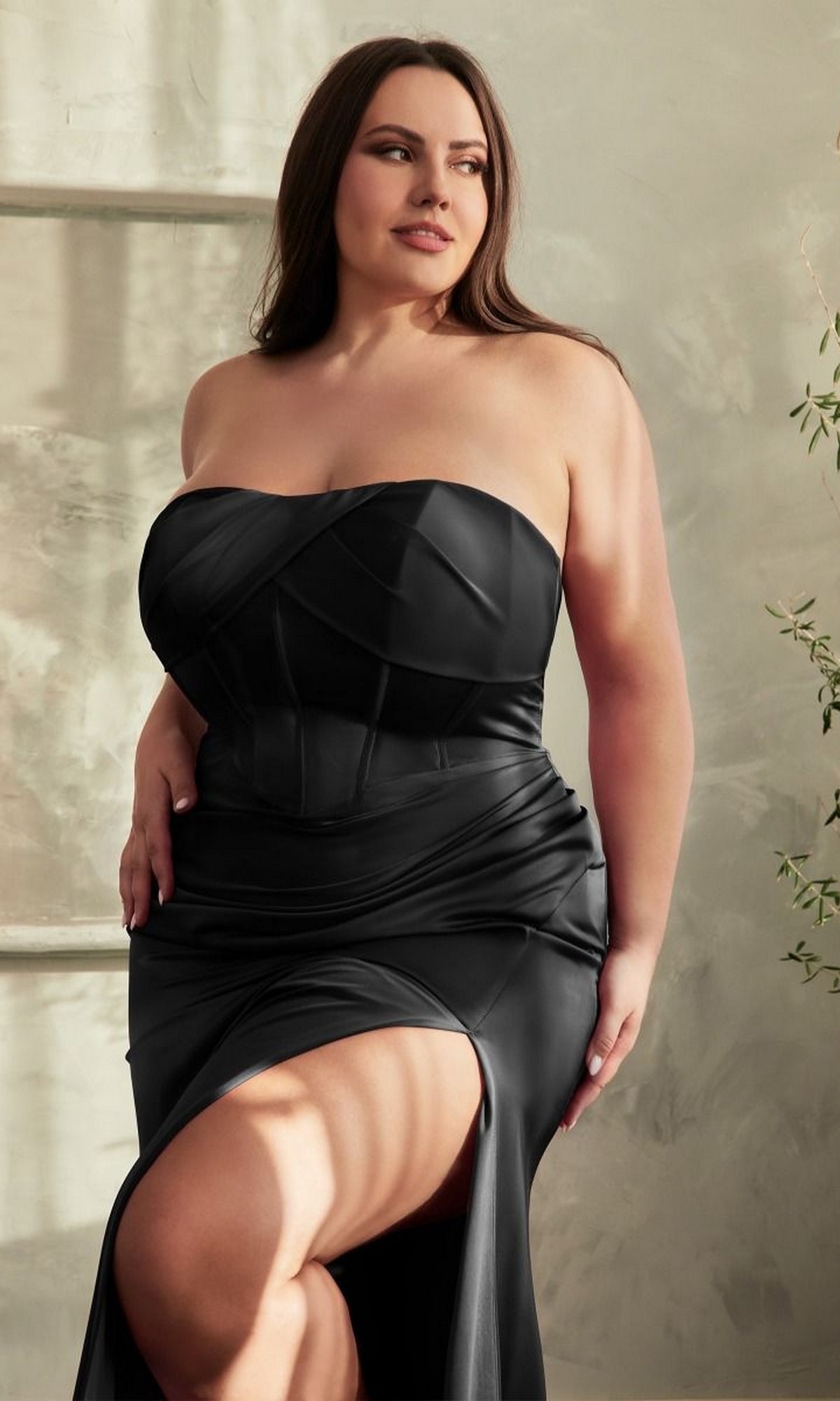 A woman wears the Lace-Up Plus-Size Strapless Long Prom Dress CD326C, featuring a corset-style bodice and thigh-high slit, as she stands indoors looking to the side.
