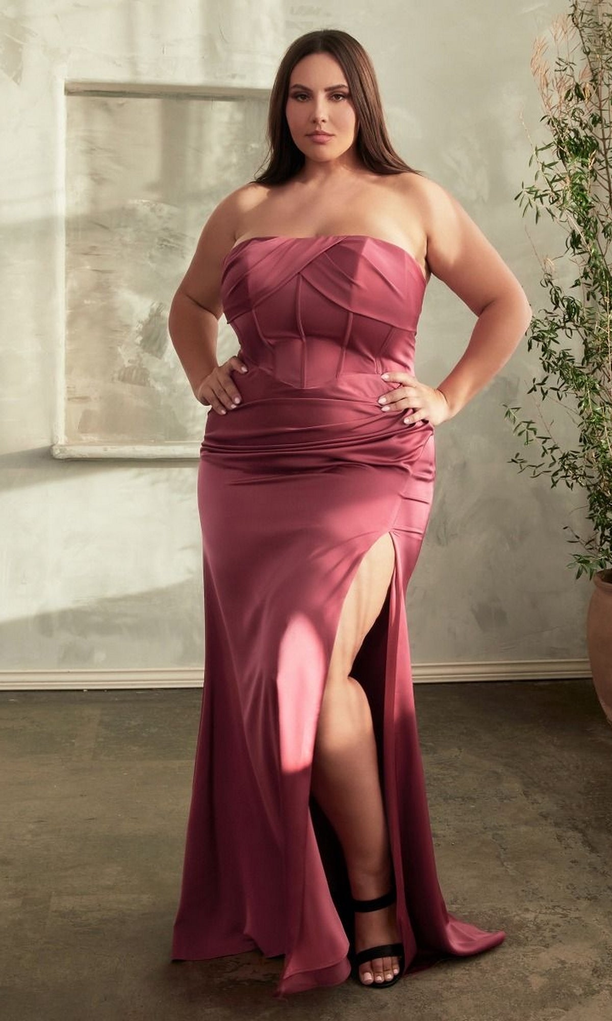 A woman wears the Lace-Up Plus-Size Strapless Long Prom Dress CD326C, featuring a corset-style bodice and thigh-high slit, while standing indoors on a concrete floor beside a potted plant.