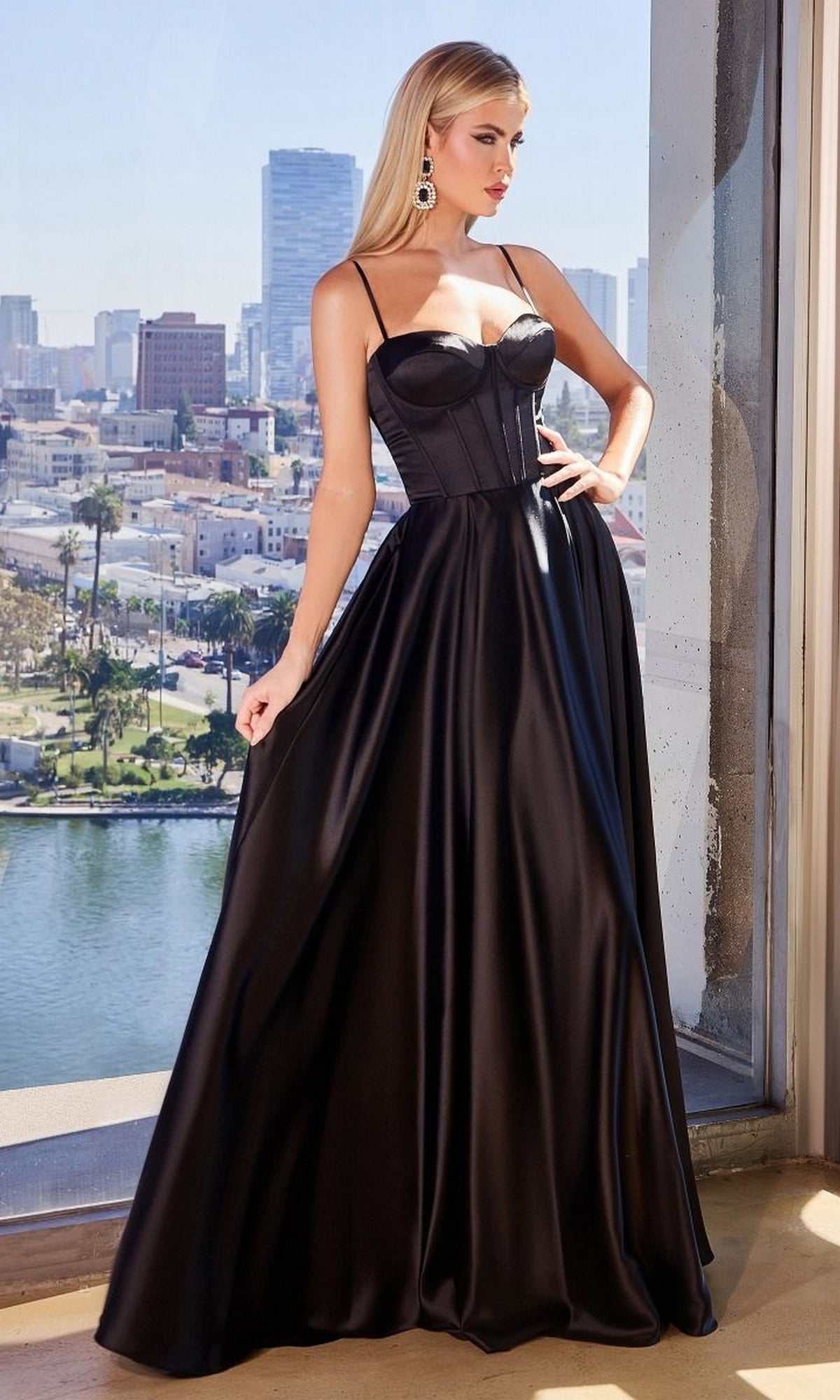 A woman in the Ladivine CD337 Long Sweetheart A-Line Prom Dress, featuring thin straps and a corset bodice, stands by a window overlooking the city during daylight.