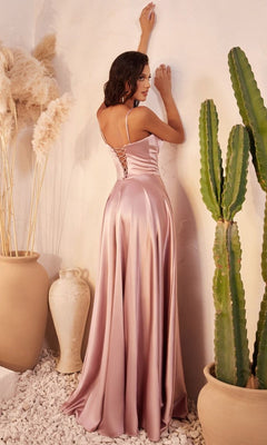 A woman wearing the Long Sweetheart A-Line Prom Dress: Ladivine CD337, a light pink satin gown with a lace-up back, stands facing a wall amid potted plants and white stone decor.