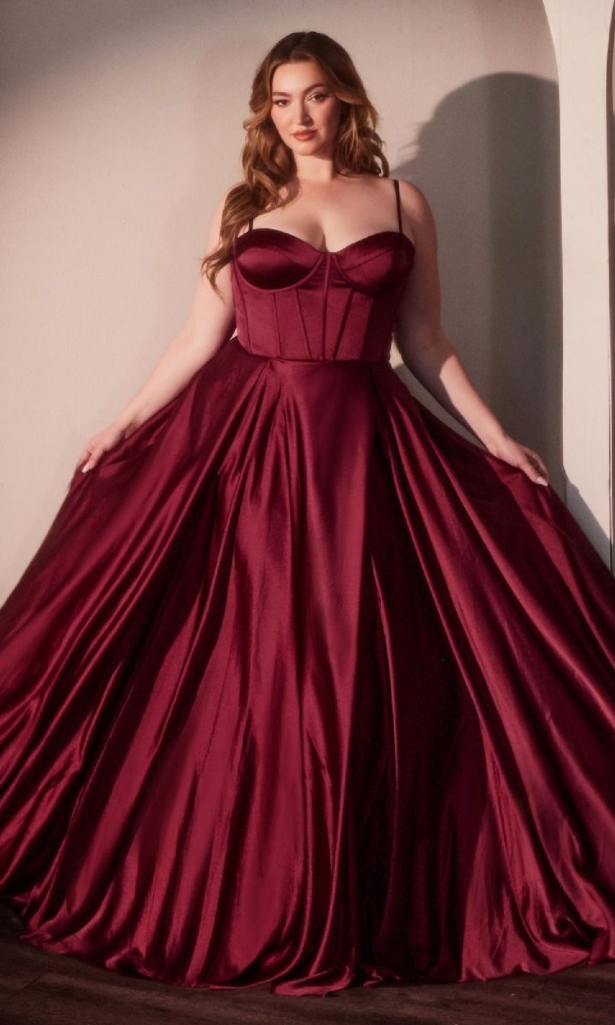 A woman wears the Ladivine CD337C Plus-Size Long A-Line Prom Dress, featuring a fitted bodice and full skirt in burgundy satin, as she stands against a pale wall holding out the sides of her gown.