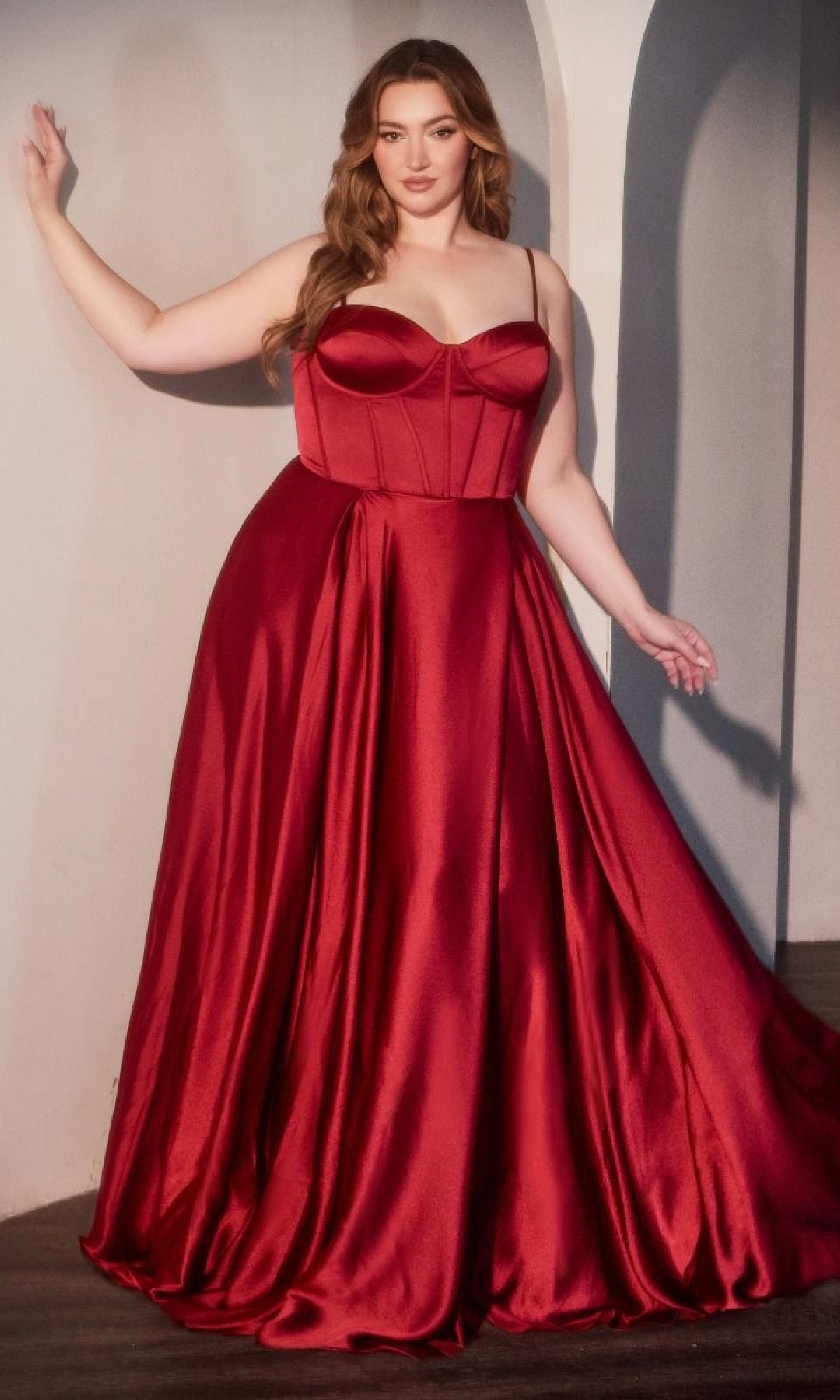 A woman poses indoors in the Ladivine CD337C, a plus-size long A-line prom dress featuring a sleeveless, fitted bodice and full skirt in striking red satin.