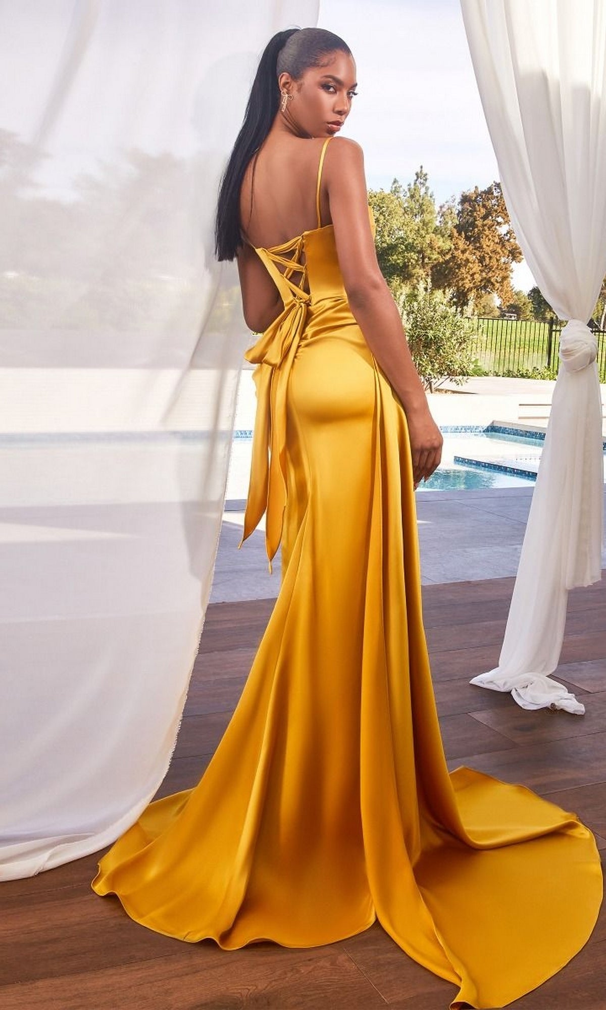 A woman in the Beaded Neckline Long Side-Drape Prom Dress CD340, featuring a fitted silhouette and lace-up back with a long train, stands on a wooden deck by the pool, gazing over her shoulder.