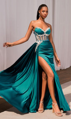 A woman in the Ladivine CD343 Sheer Corset Long Satin Prom Dress in teal, with a strapless neckline, thigh-high slit, and white lace accents, stands on a wooden floor before white draped curtains.