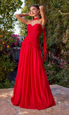 A woman stands outdoors in the Ladivine CD395, a long, strapless red prom dress with a corset bodice and matching neck scarf, surrounded by lush greenery and colorful flowers.
