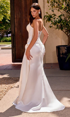 A woman is wearing the Ladivine CD636W long prom dress—a sleeveless, floor-length white satin gown featuring a strapless scoop neckline and lace-up back detail—while standing outdoors near a door and large potted plant.