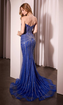 A woman in the Royal Blue Long Beaded Prom Dress: Ladivine CD785B, featuring a strapless fitted floor-length design with silver accents, stands facing away in a softly lit room with white curtains.