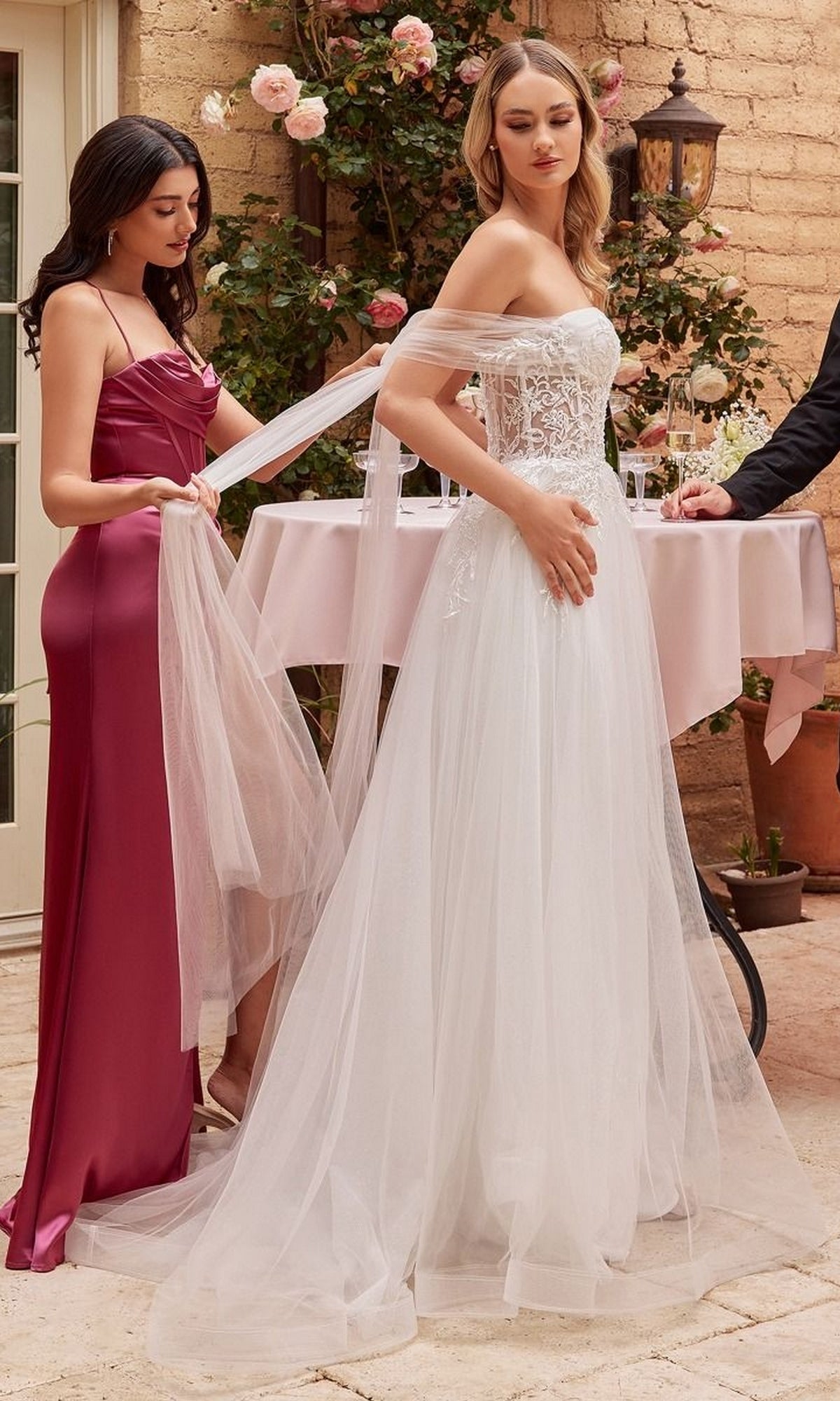 A woman wears the Off White Long Halter Wedding Dress CD827W as another in a burgundy gown adjusts her veil; they are outdoors by a table with drinks and flowers.