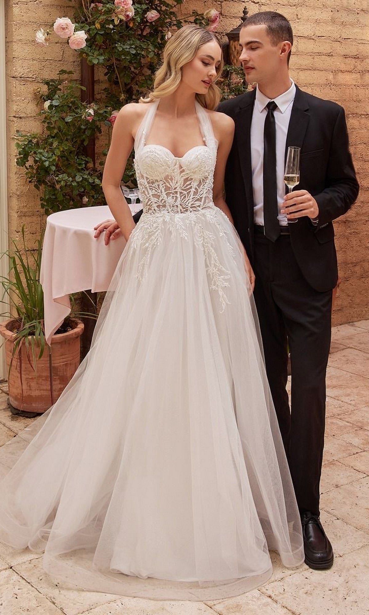 A woman wearing the Off White Long Halter Wedding Dress CD827W stands beside a man in a black suit and tie, holding a champagne glass, in an outdoor garden setting.
