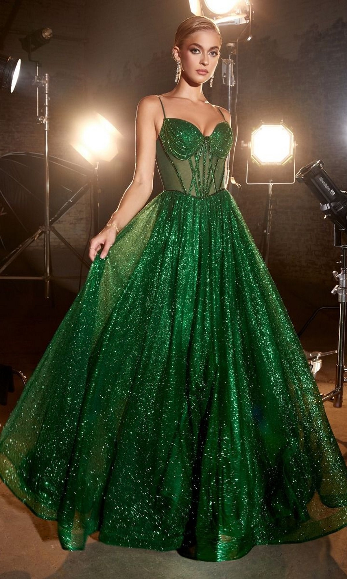 A woman stands in a studio, surrounded by professional lighting, wearing the Sheer-Bodice Long A-Line Shimmer Prom Dress CD832 with its sparkling green fitted corset bodice.
