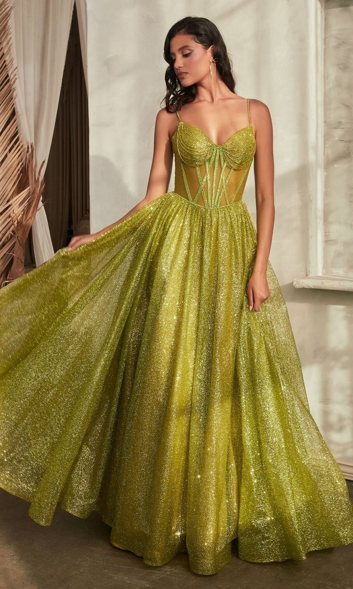 A woman indoors wears the Sheer-Bodice Long A-Line Shimmer Prom Dress CD832, a floor-length sparkling green gown with a structured bodice and spaghetti straps, elegantly holding out part of its skirt.