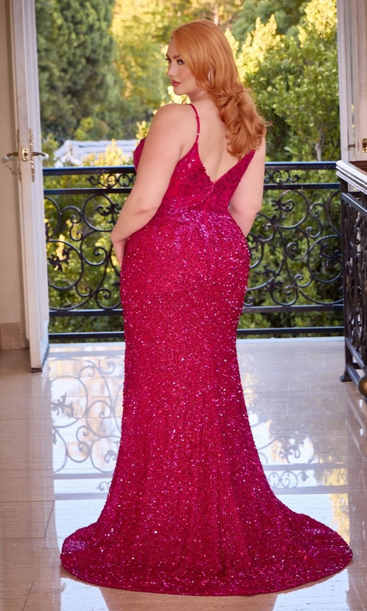 A red-haired woman stands indoors before open doors, wearing the V-Neck Plus-Size Long Sequin Prom Dress CD840C in pink. The floor-length gown features a low back and train. Trees and a balcony can be seen outside.