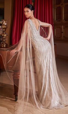 A woman poses in the Long Platinum Formal Dress CD865, featuring sheer cape sleeves and a deep V-back, by a wooden table with red curtains in the background.