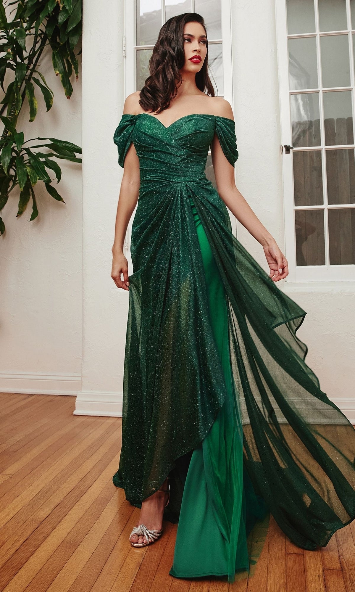 A woman stands indoors wearing the Wrap-Waist Long Glitter Prom Dress CD878 in dark green with a sheer overlay, paired with silver open-toe heels.