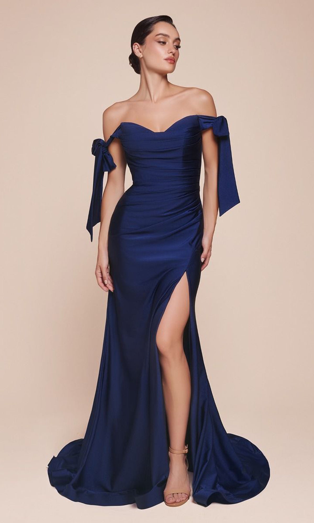 A woman in the navy blue Off-Shoulder Ruched Prom Dress: Ladivine CD943, featuring a fitted silhouette, side bows, and a thigh-high slit, stands against a plain beige background.