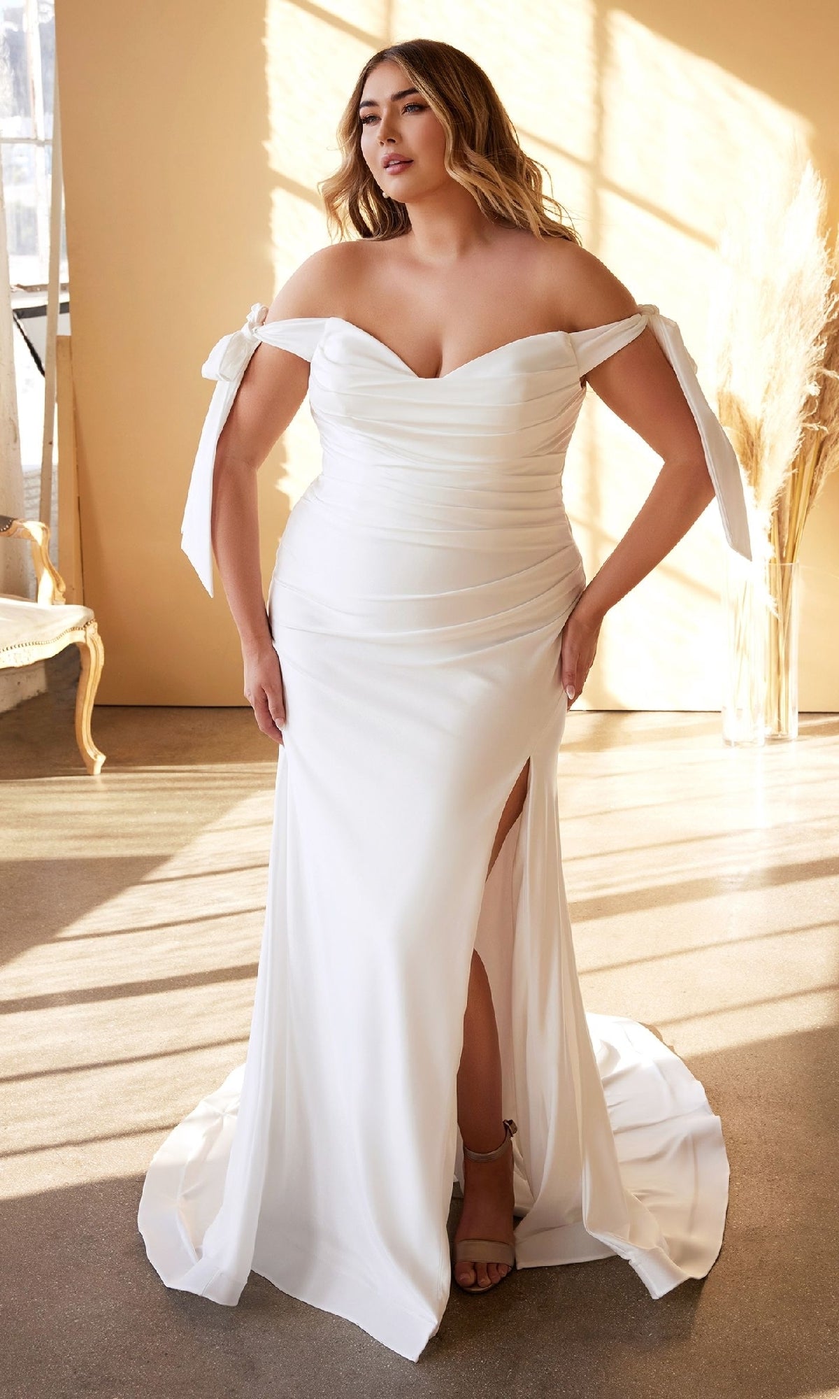 A woman stands indoors, beautifully lit by natural light, wearing the Ladivine CD944WC—a long plus-size bridal dress featuring an off-shoulder neckline and a thigh-high slit.