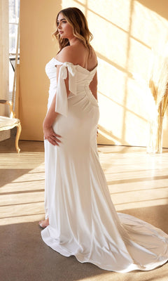 A woman wears the Ladivine CD944WC Long Plus-Size Bridal Dress, an off-shoulder, floor-length white satin jersey gown with a train. She stands indoors amid neutral decor and sunlight, looking back over her shoulder.