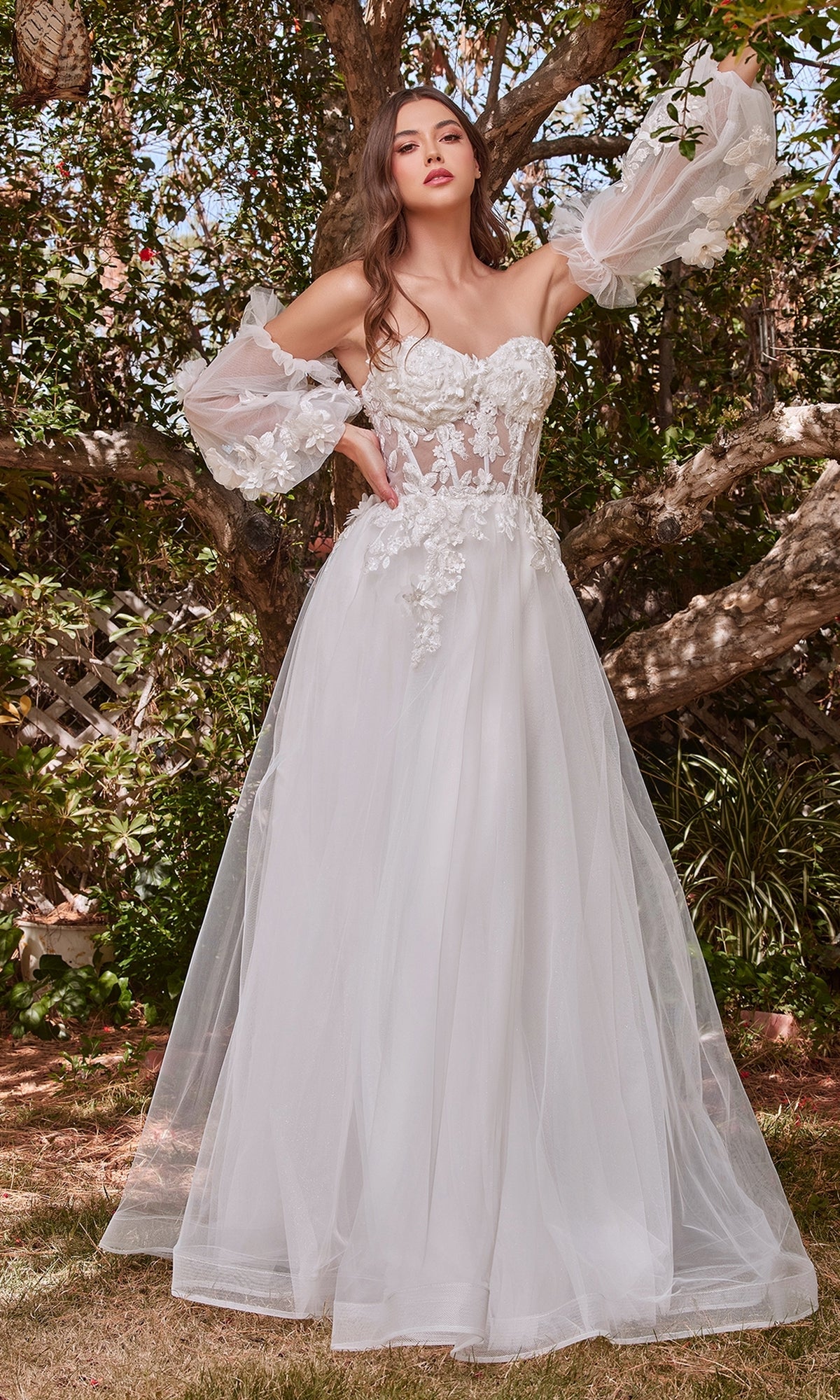 A woman stands outdoors in front of trees wearing the Puff-Sleeve Strapless Long White Bridal Gown CD962W, featuring sheer puffed sleeves and floral lace details.