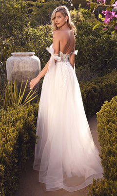 A woman in the Ladivine CD964W long bridal dress with an off-white tulle A-line skirt stands on a garden path, looking over her shoulder amid lush greenery and blooming flowers.