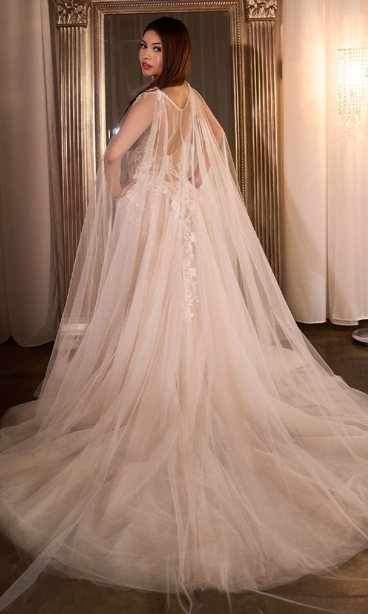 A woman wearing the Ladivine CDS476W long wedding dress with a flowing floral lace design and sheer train poses with her back to the camera in front of a large ornate mirror.