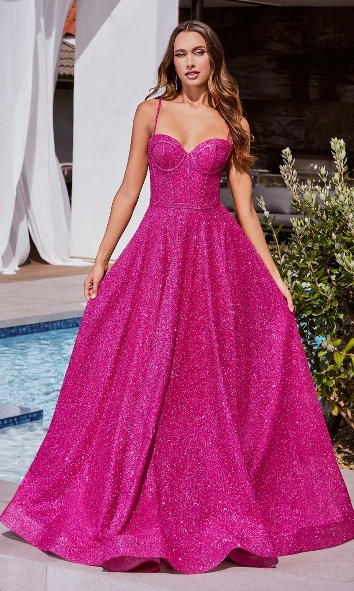 A woman stands outdoors by a pool, wearing the Long Glitter Bustier-Bodice Prom Ball Gown CDS483—a sparkling hot pink dress with thin straps and a fitted corset-style bodice—holding out the sides of the gown.