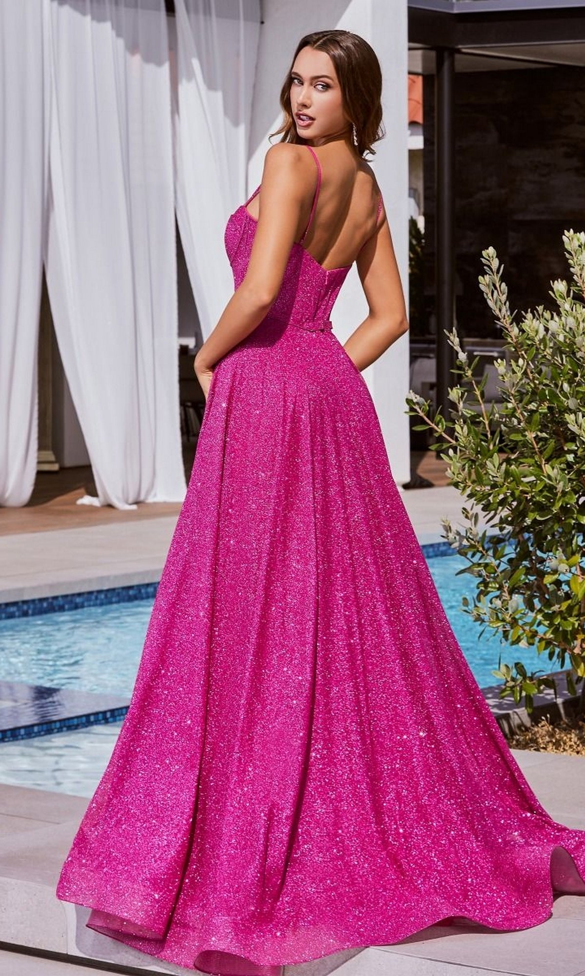A woman stands by the pool in the Long Glitter Bustier-Bodice Prom Ball Gown CDS483, a floor-length hot pink dress with sparkly fabric, a corset-style bodice, and thin straps, looking over her shoulder.