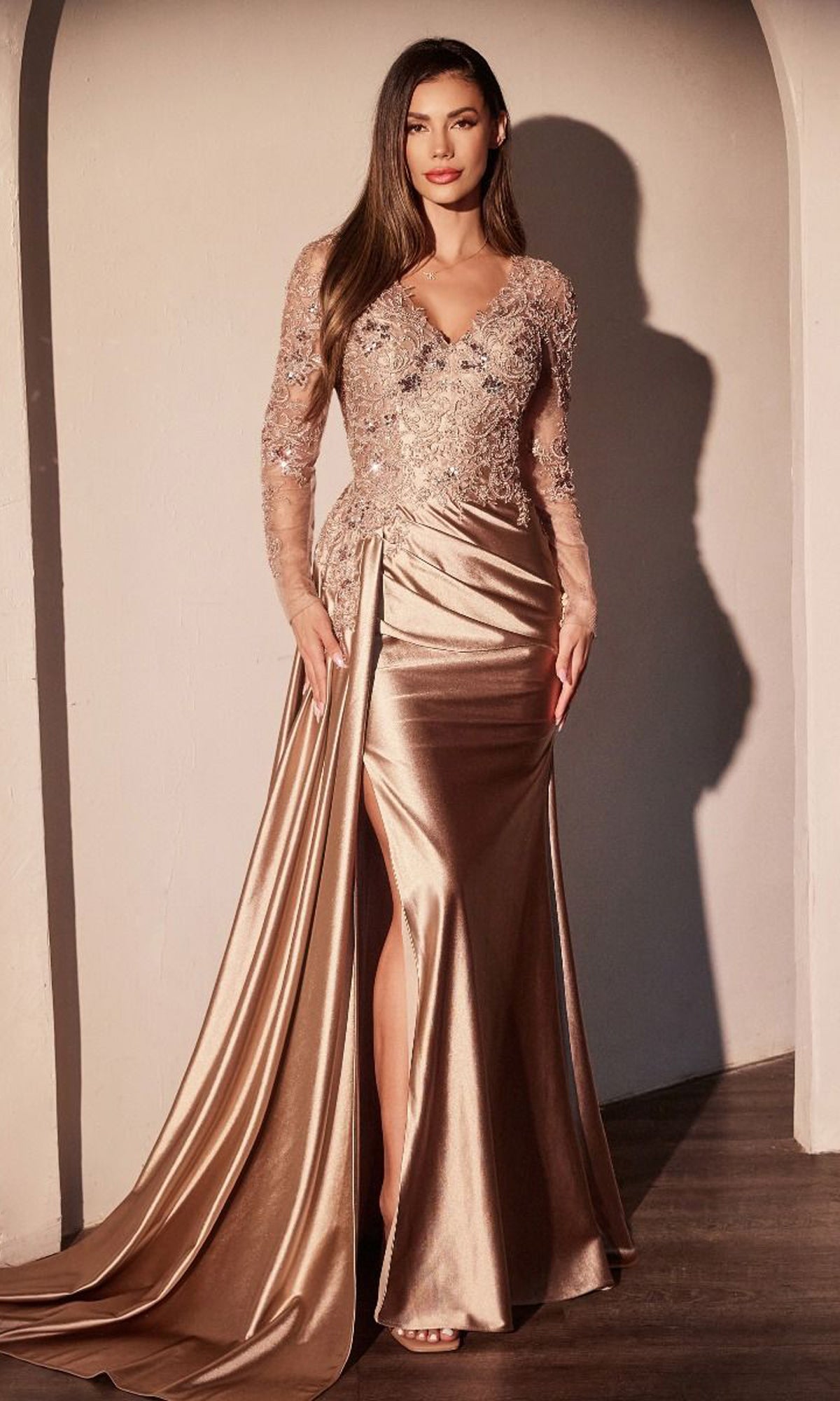 A woman stands indoors wearing the Ladivine CDS506, a long sleeve formal dress featuring gold satin, a thigh-high slit, and an embellished lace bodice.