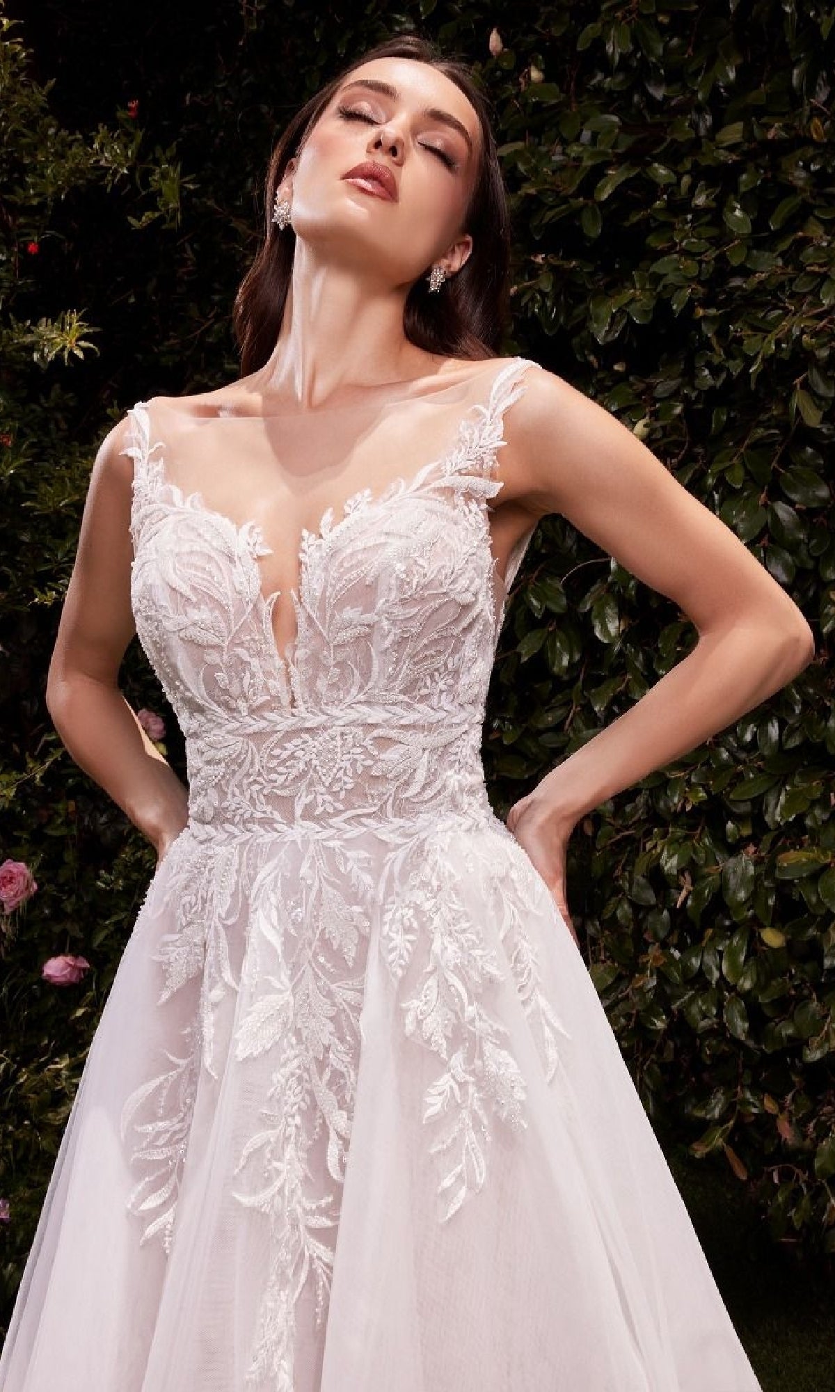 A woman stands outdoors before leafy greenery wearing the Ladivine CDS511W, a long white wedding dress with sheer straps and a lace-embroidered bodice.