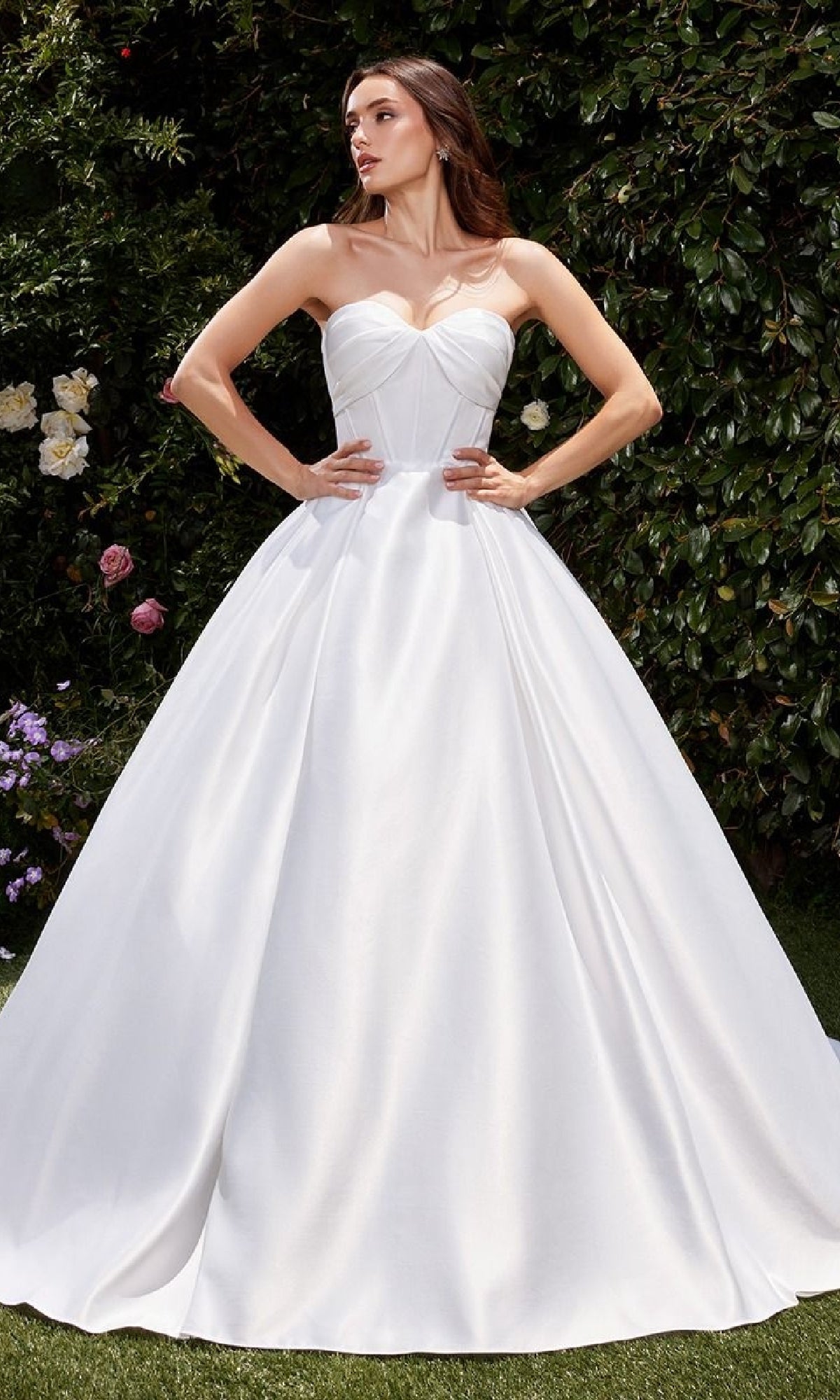 A woman wearing the Ladivine CDS512W White Satin Formal Ball Gown stands outdoors amid lush green foliage and blooming flowers.
