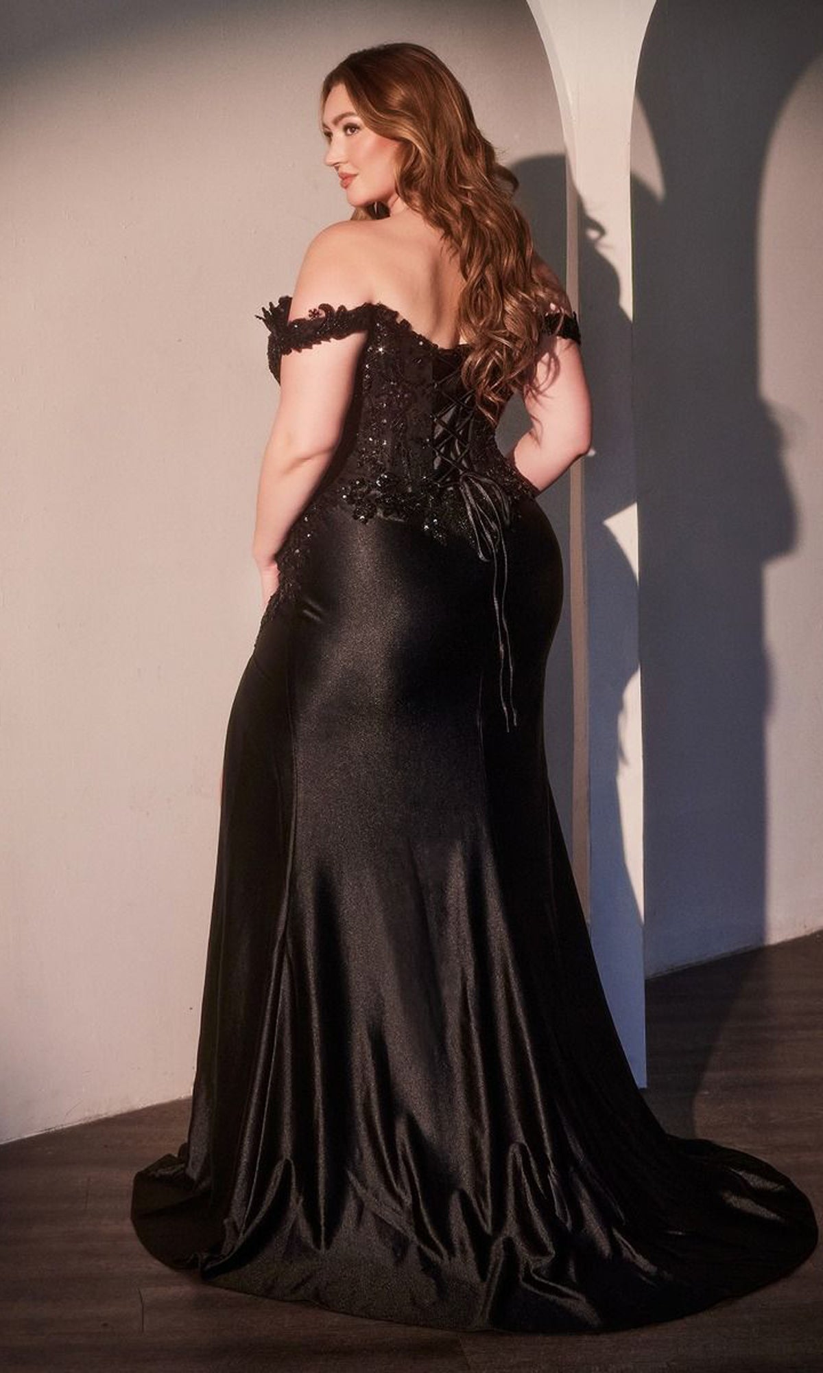 A woman with long wavy hair wears the Ladivine CDS537C Plus-Size Off-Shoulder Prom Gown with a lace-up back, standing in a room with white walls and wooden floors.