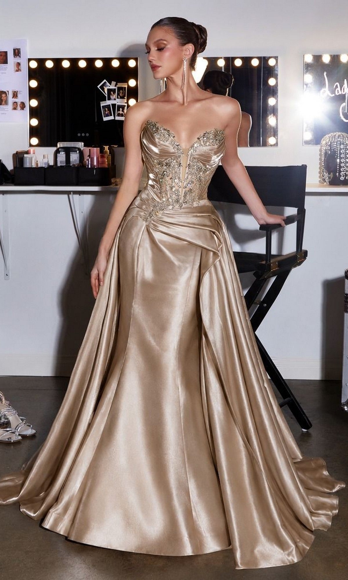 A woman stands in a dressing room beside a director’s chair and lit vanity mirror, wearing the Ladivine CDS568 Long Prom Dress in Mocha Gold, featuring a strapless fitted bodice and flared organza skirt.