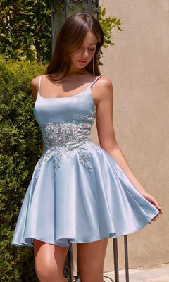 A young woman stands outdoors near greenery and a wall, wearing the Ladivine CH2613 short homecoming dress in blue—a sleeveless, light A-line style with lace detailing.