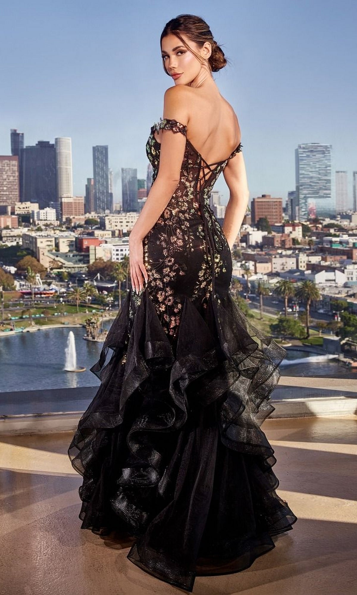 A woman in the Ladivine CM354 Black Floral Mermaid Prom Gown, featuring an off-the-shoulder neckline and lace-up back, stands on a rooftop overlooking the city skyline during the day.