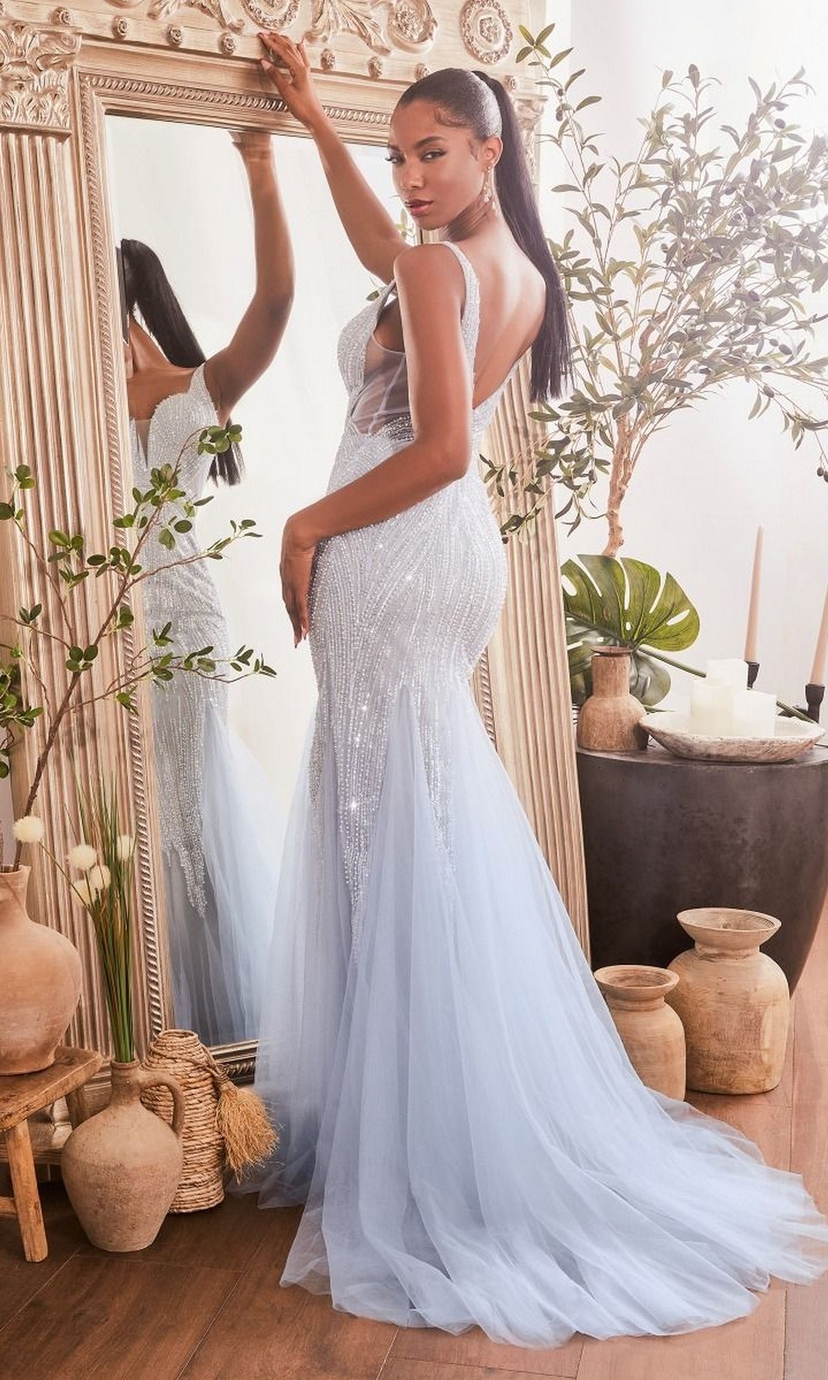 A woman poses in front of a large ornate mirror, surrounded by decorative plants and pottery, wearing the Ladivine CR874 Sheer-Sides Long Beaded Prom Dress—a light blue, sparkly evening gown—in a well-lit room.