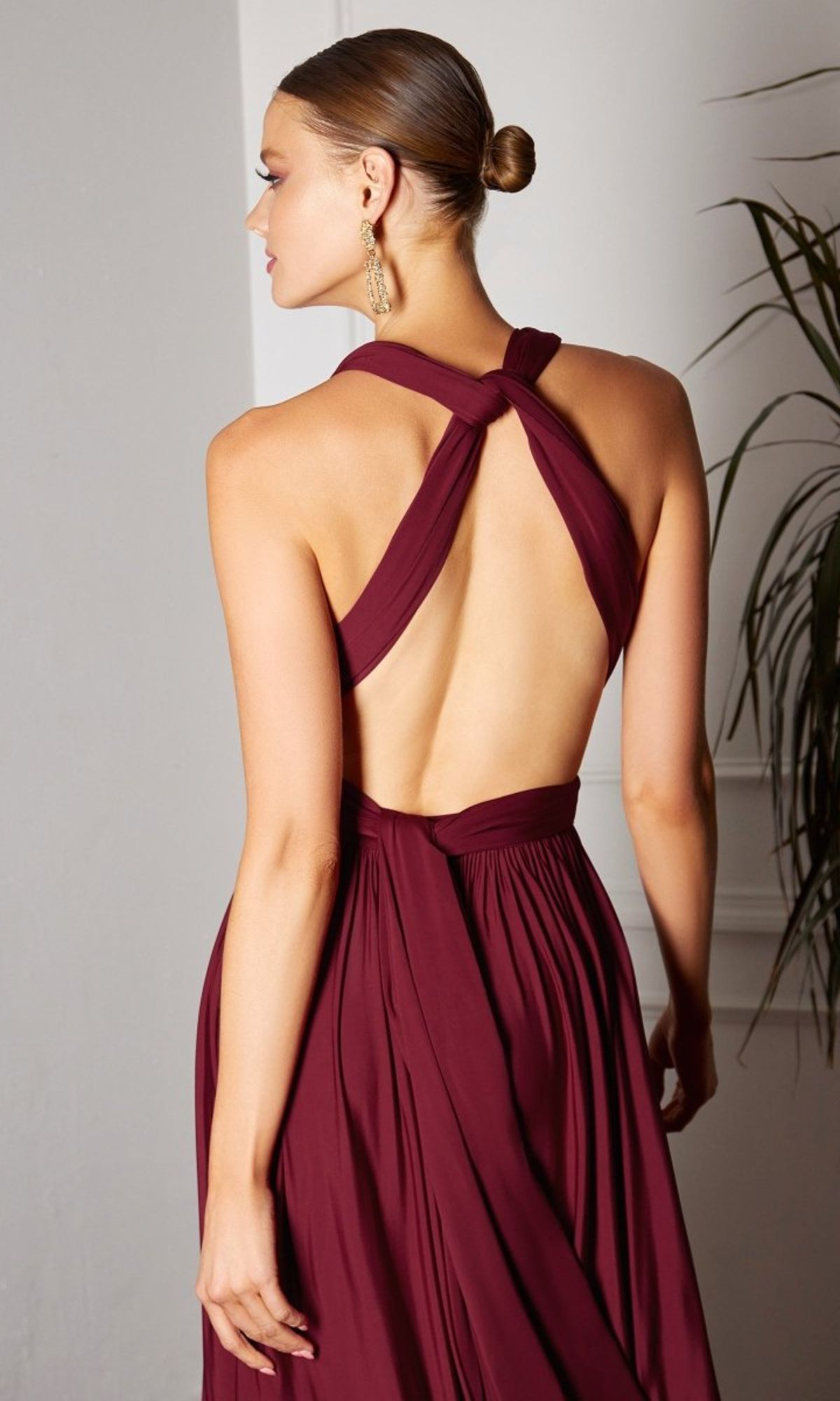A woman wears the Amoris CV01 Multi-Wrap Long A-Line Prom Dress in maroon, featuring a halter neck and open back, as she stands indoors facing away from the camera.