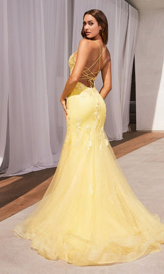 A woman in the Long Pastel Floral Mermaid Prom Dress D145, featuring a yellow hue, open back with thin crisscross straps, and floral embellishments, stands indoors near white curtains on a light floor.