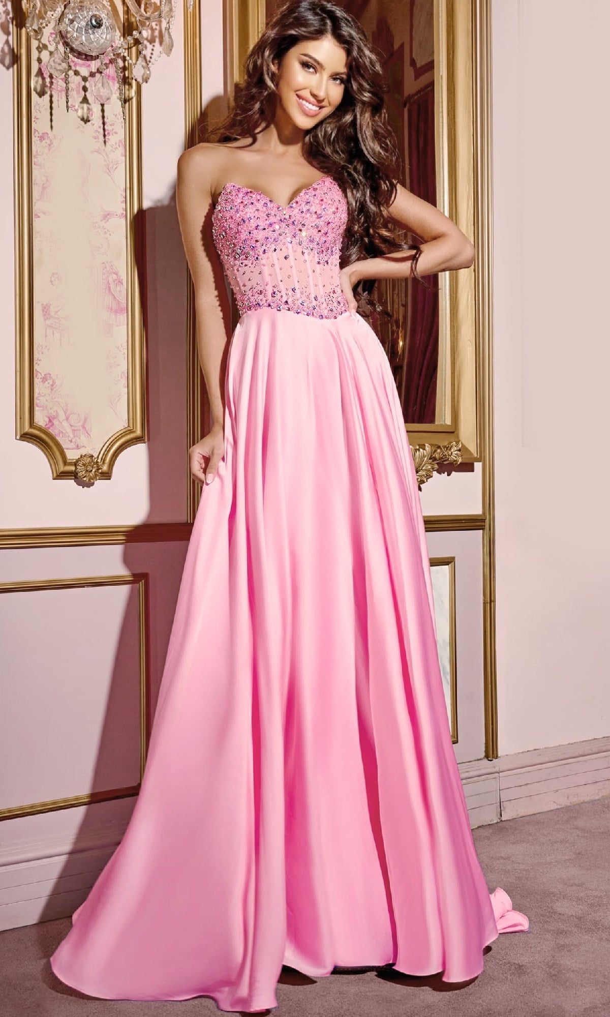 A woman models the Jovani D6021 Pink Long Prom Dress, featuring a strapless beaded corset bodice and flowing A-line skirt, as she stands indoors by an ornate wall.