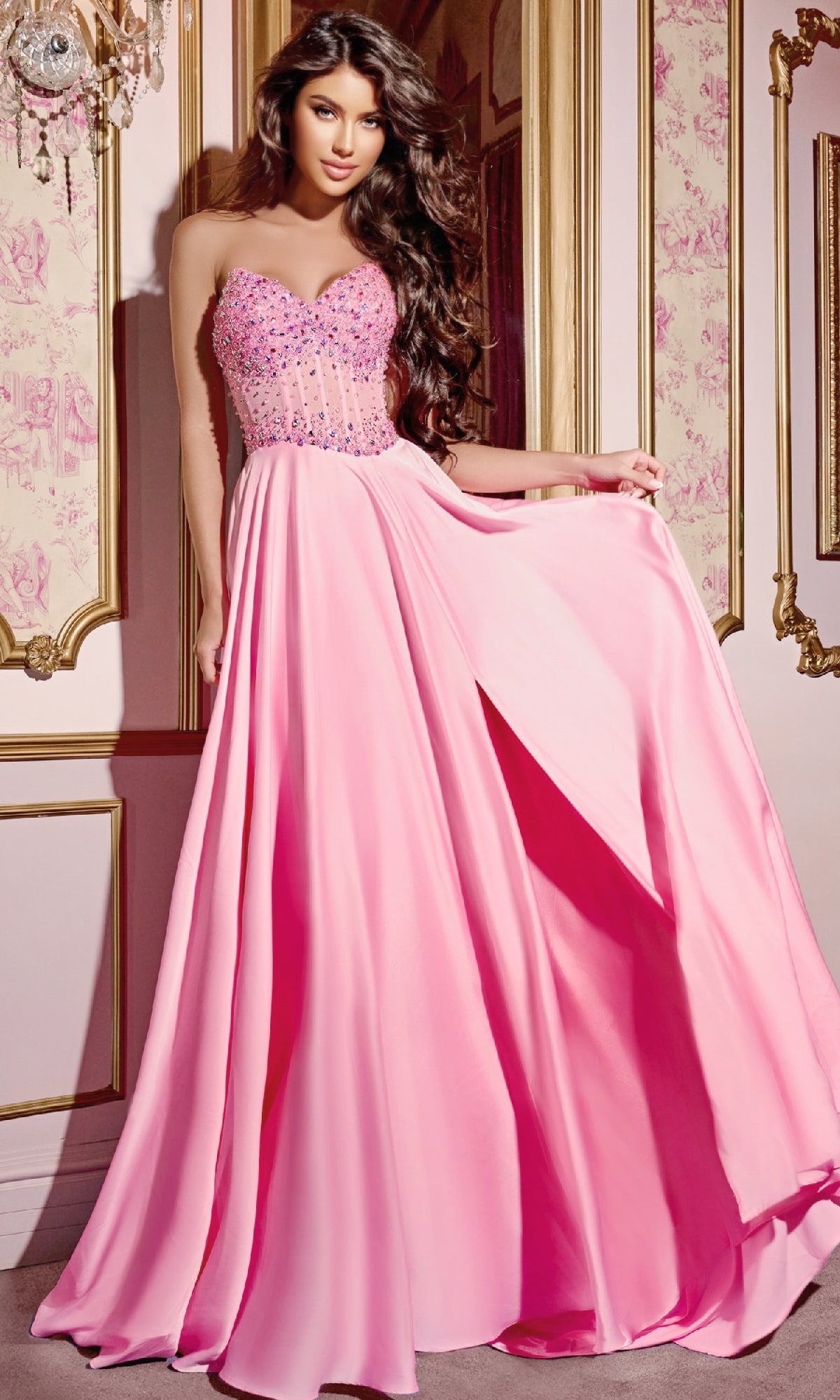 A woman wearing the Pink Long Prom Dress: Jovani D6021, with a strapless beaded corset bodice and flowing A-line skirt, poses gracefully in an ornate gold-trimmed room adorned with elegant floral wallpaper.