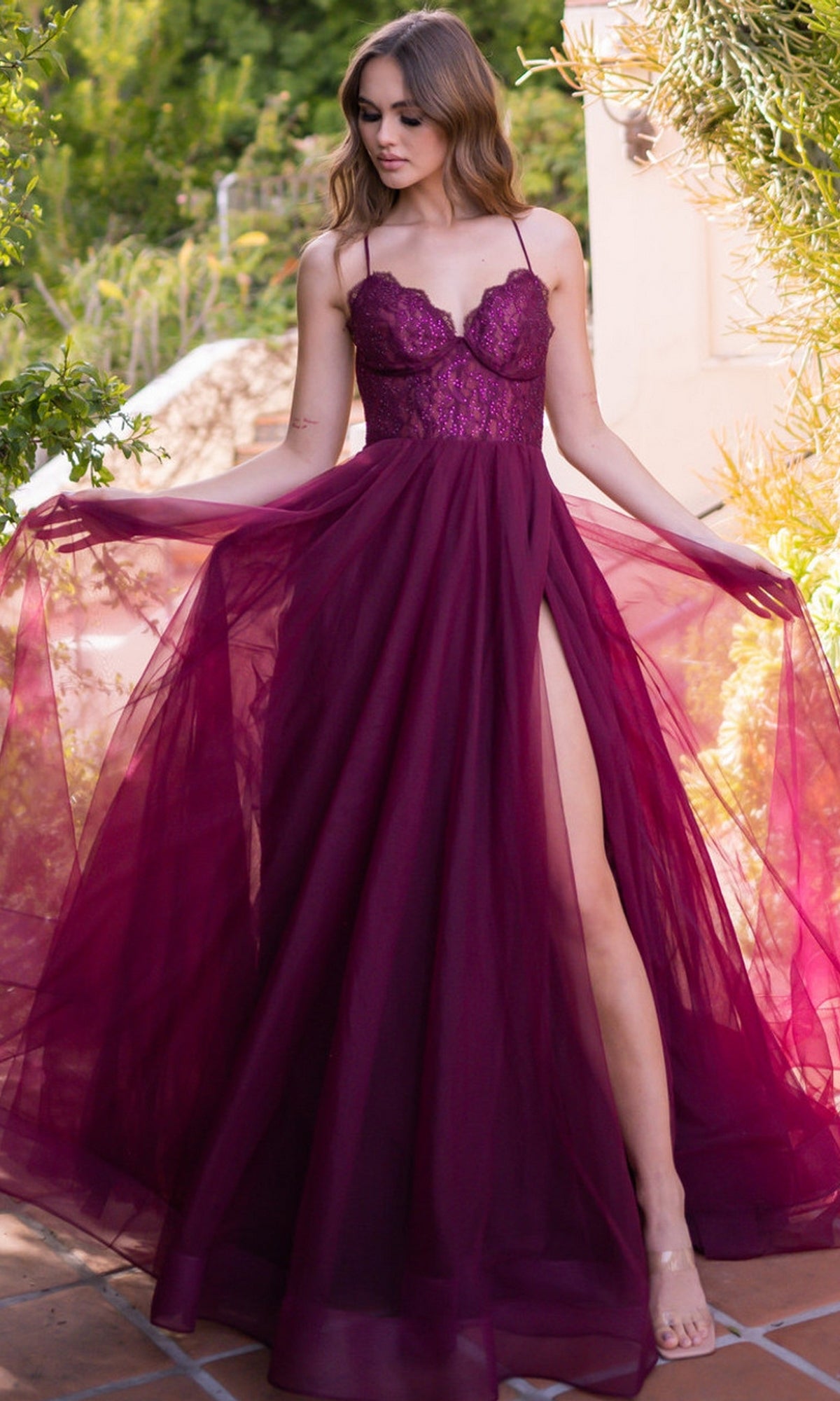 Woman wearing the Velvi Long Sweetheart A-Line Prom Dress Dani, a sleeveless, deep magenta lace gown with a tulle skirt, poses outdoors and holds the sides of her formal dress.